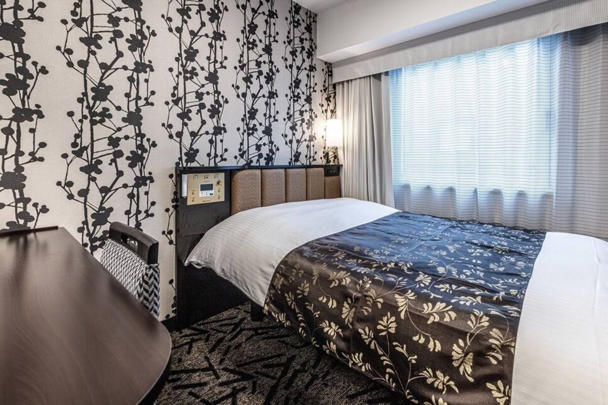 A hotel room with a bed, a desk, a nightstand, and a window with curtains.