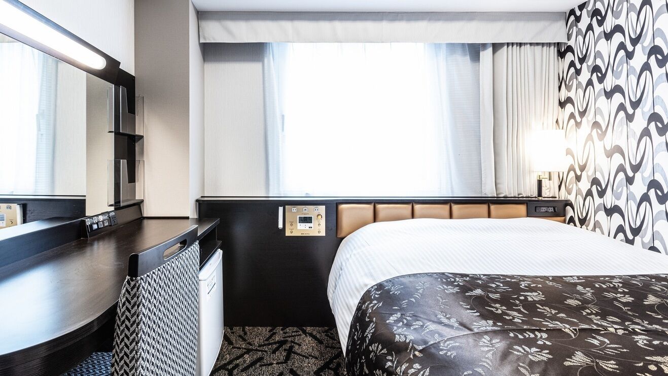 A hotel room with a bed, a desk, a mirror, and a window with curtains.