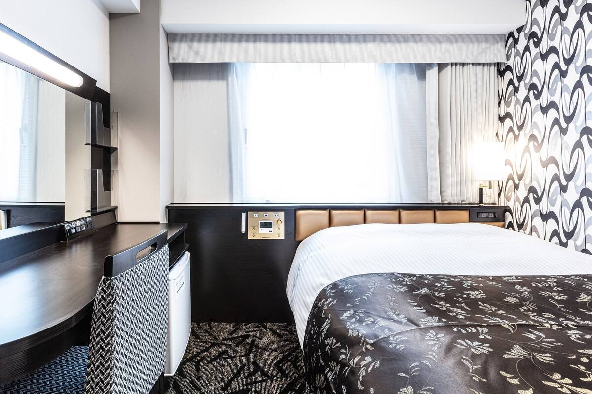 A hotel room with a bed, a desk, a chair, a mirror, a window with curtains, and a wall-mounted control panel.