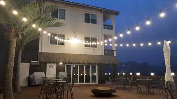Un edificio de dos pisos con un patio techado de paja, luces colgantes y mobiliario exterior.