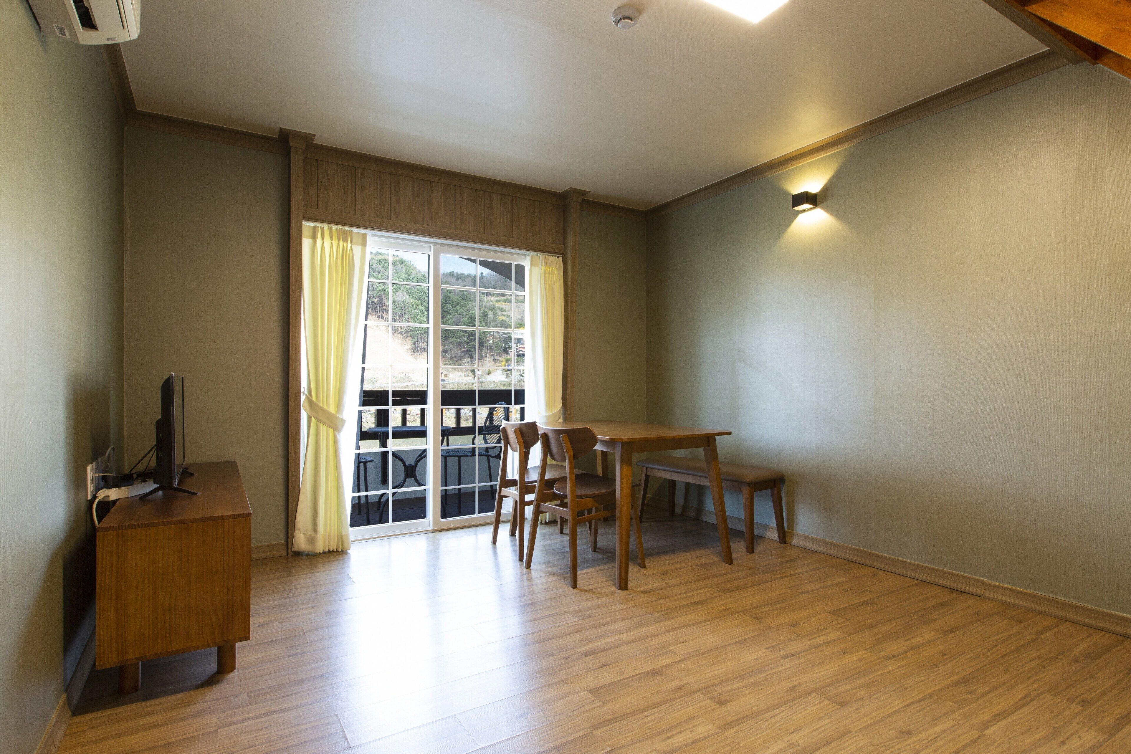 A room with a wooden desk, a dining table with chairs, a sliding glass door with curtains, and a ceiling-mounted light fixture.