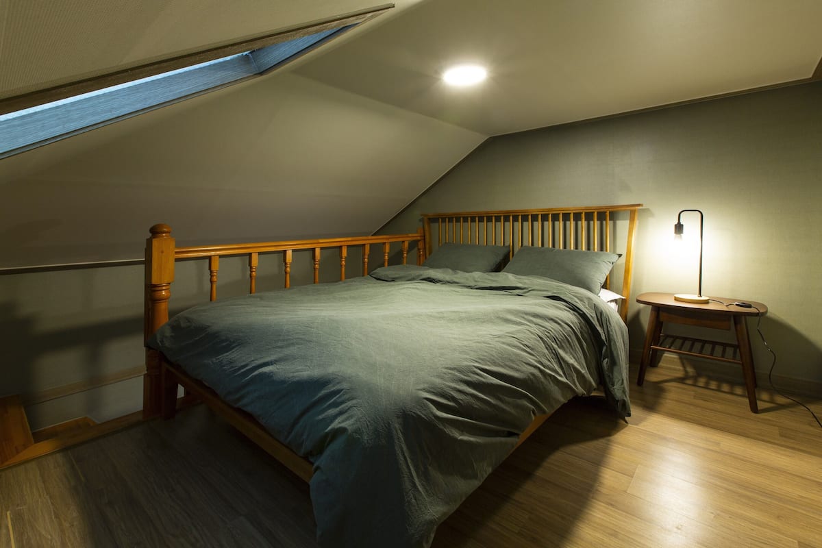 Une chambre avec un plafond en pente, un cadre de lit en bois, une table de chevet avec une lampe et un puits de lumière.