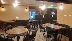 Un restaurante con mesas de madera redondas y sillas negras, una zona de bar con un letrero de neón y un refrigerador lleno de bebidas.