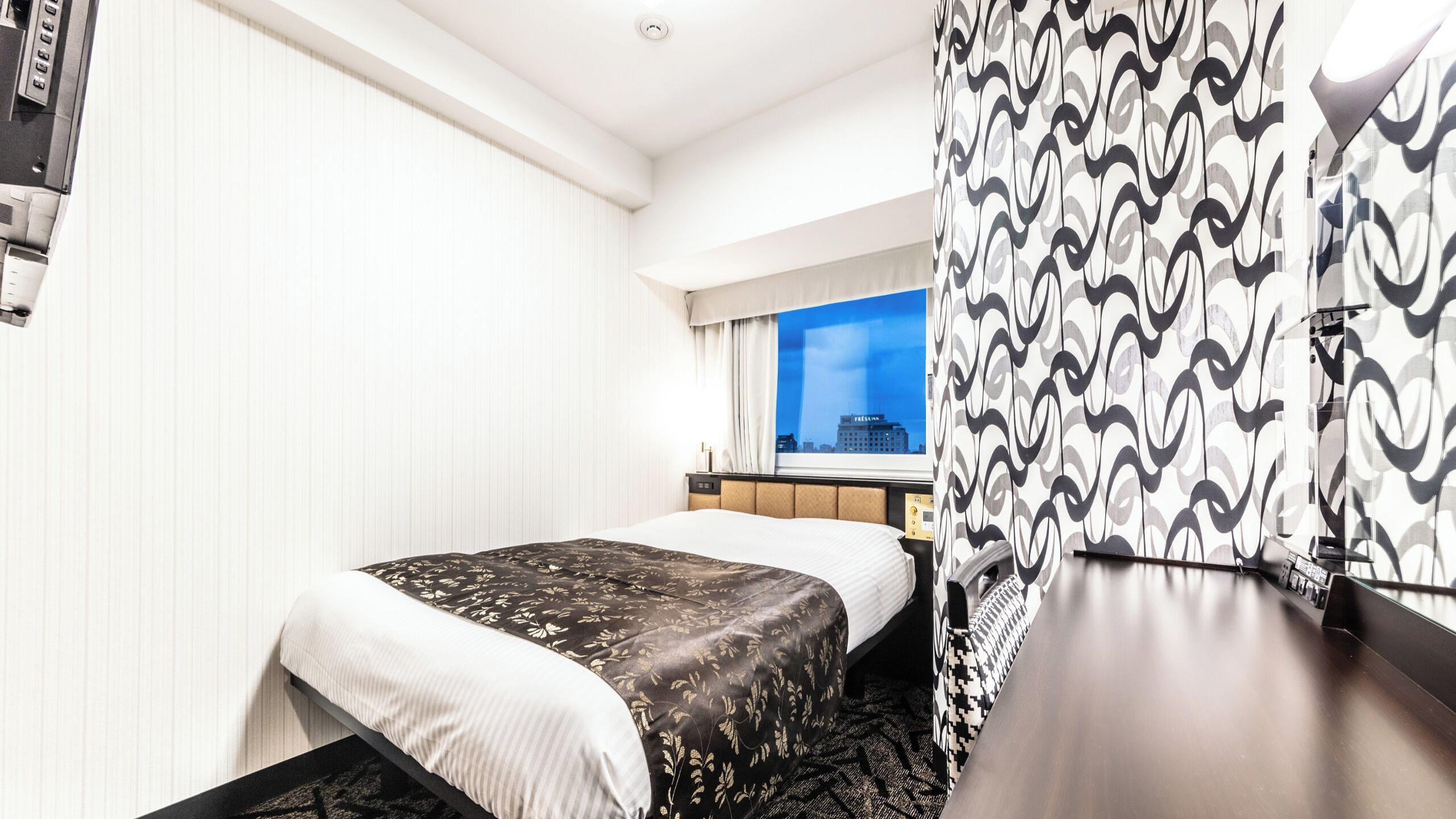 Quarto de hotel com cama, escrivaninha, cadeira, janela com vista para a cidade e cortina estampada.