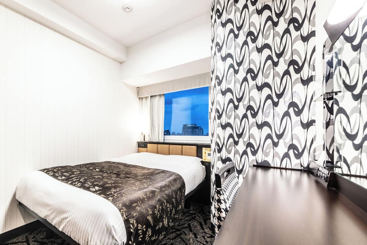 A hotel room with a bed, a desk, a chair, a window with a city view, and a patterned curtain.
