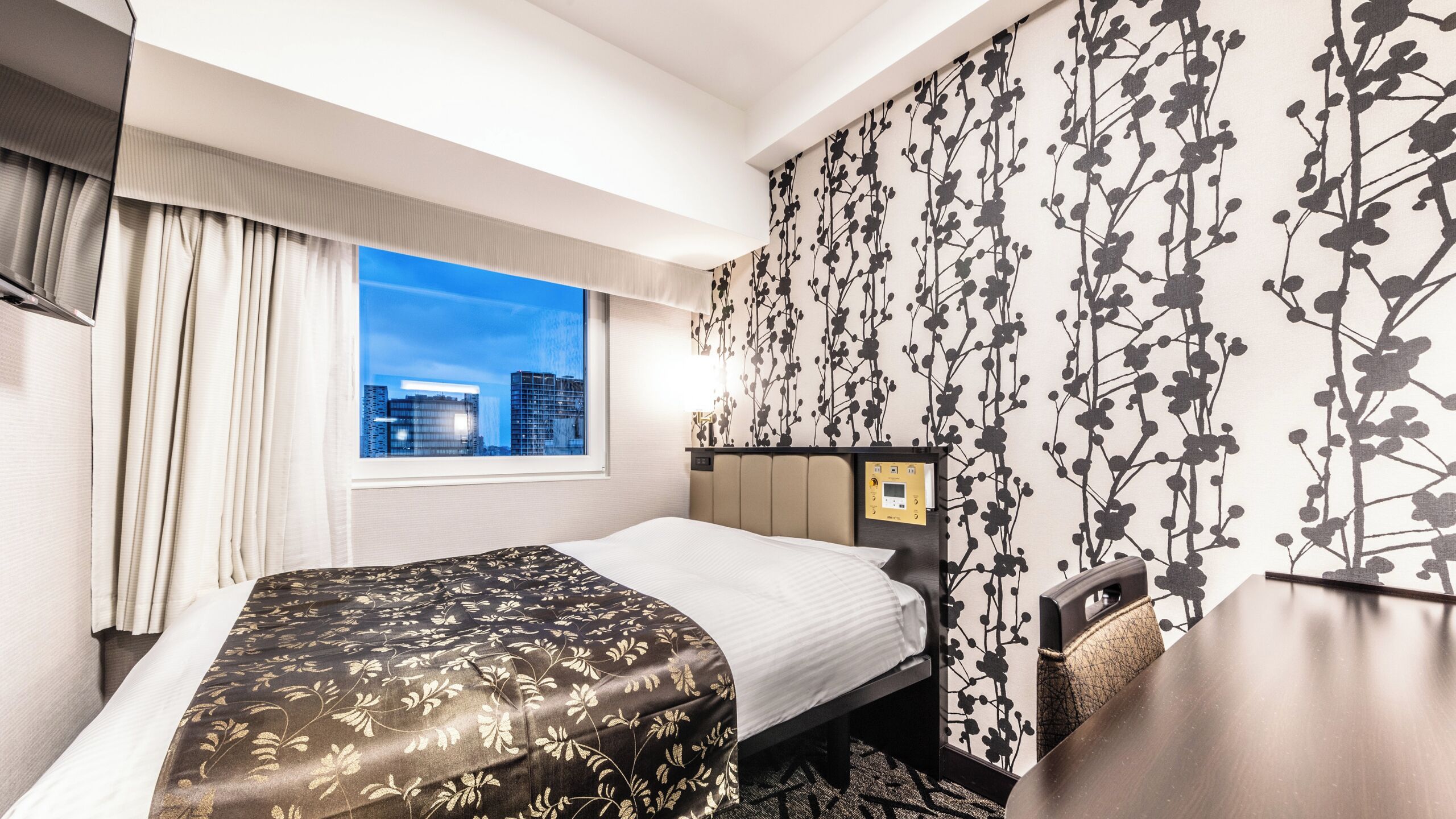 Quarto de hotel com cama, mesa de cabeceira, janela e vista da cidade, além de uma parede com padrão floral.