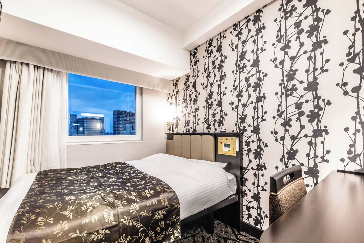 A hotel room with a bed, a nightstand, a window with a city view, and a wall with a floral pattern.