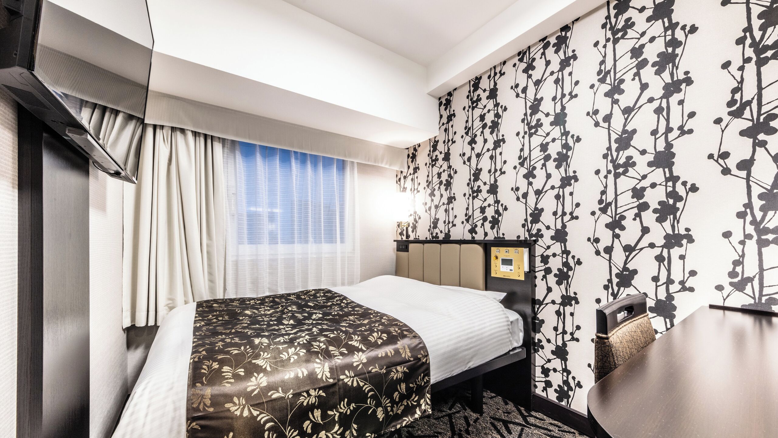 Quarto de hotel com cama, painel de controle, janela com cortinas e parede com padrão floral.