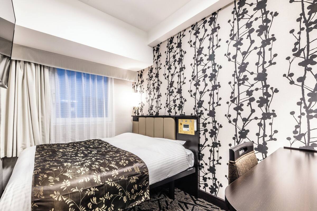 A hotel room with a bed, a control panel, a window with curtains, and a wall with a floral pattern.