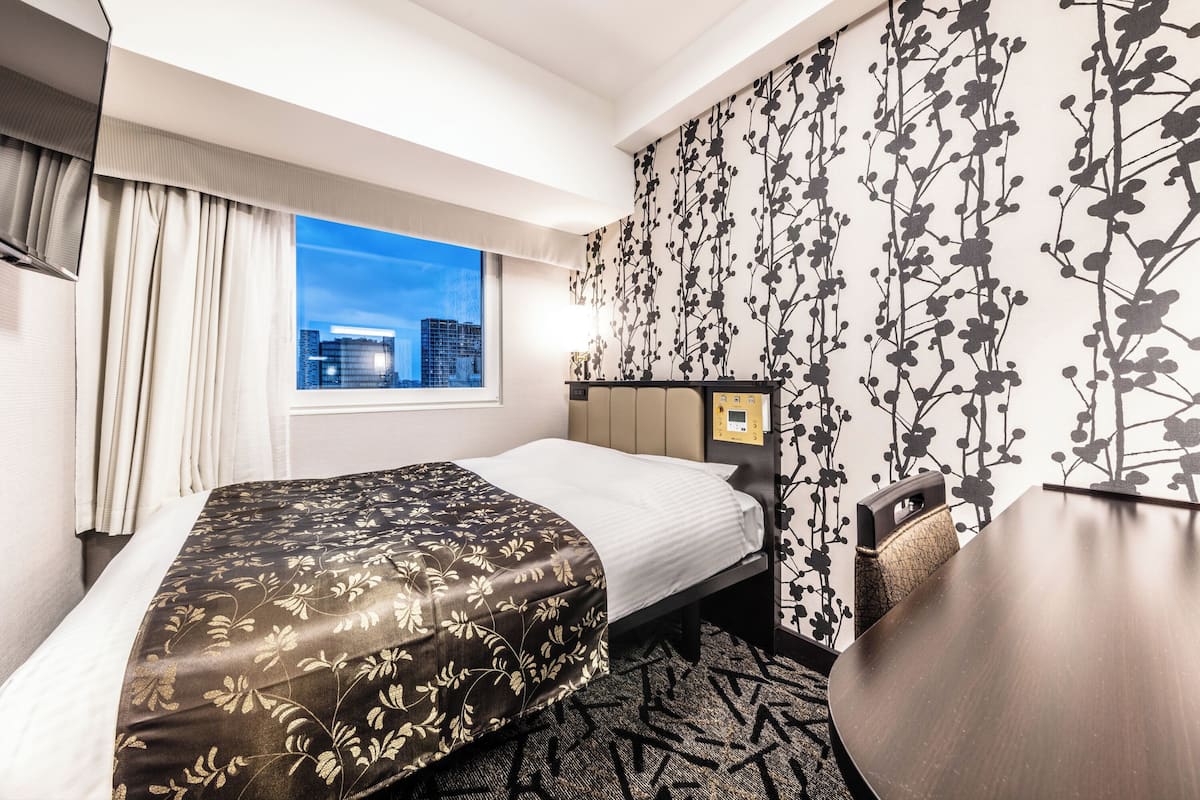 A hotel room with a bed, a desk, a view of the cityscape through the window, and a wall with a floral pattern.