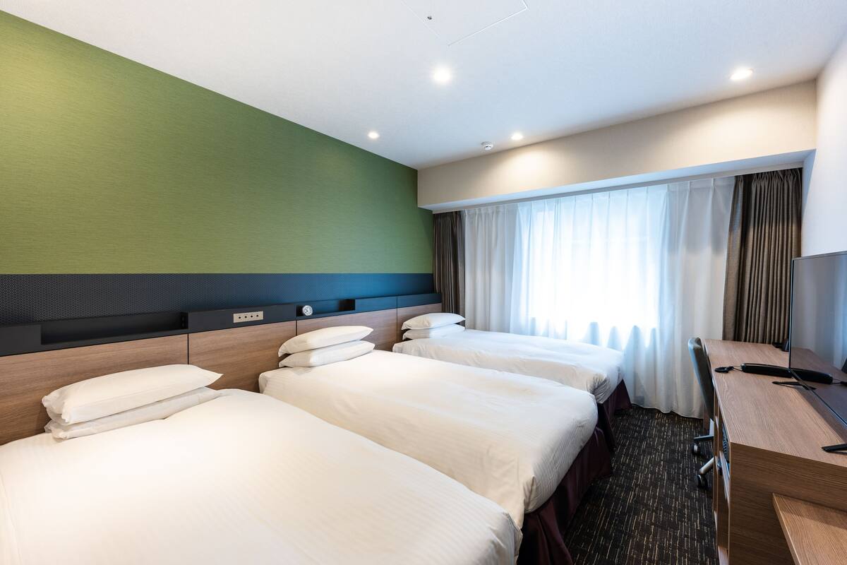A hotel room with three beds, a desk, and a large window with curtains.