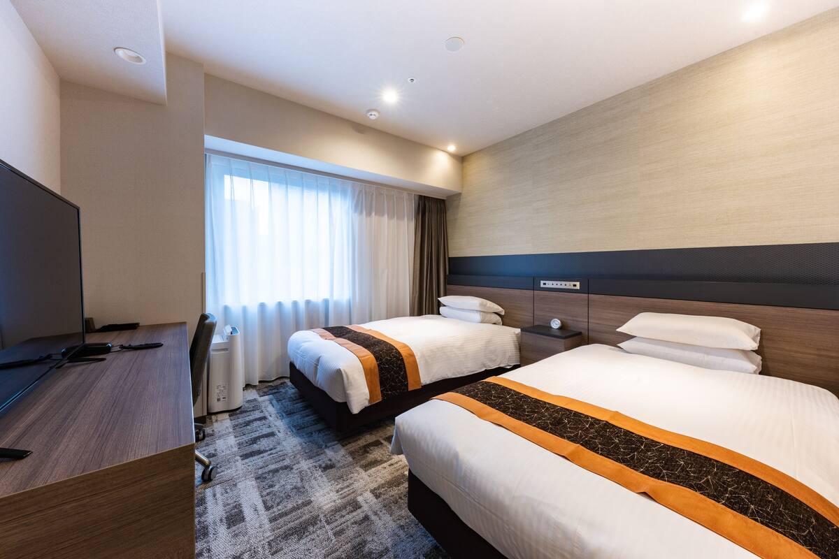 A hotel room with two beds, a desk, a television, and a window with curtains.