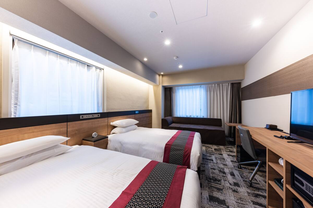 A hotel room with two beds, a desk, a TV, and a window with curtains.