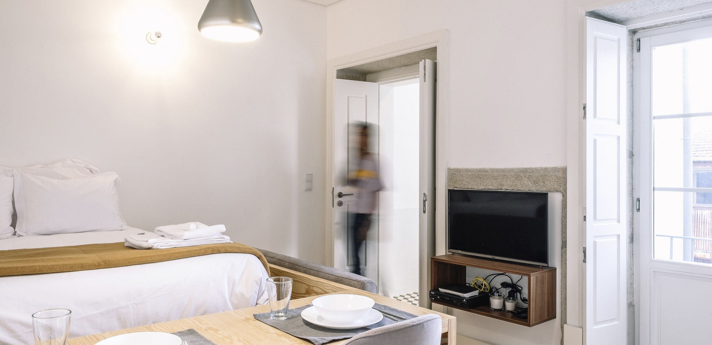 Ein Hotelzimmer mit einem Bett, einem Tisch mit Glas und Tellern, einem Fernseher auf einem Holzboden und einer Tür zu einem anderen Raum.
