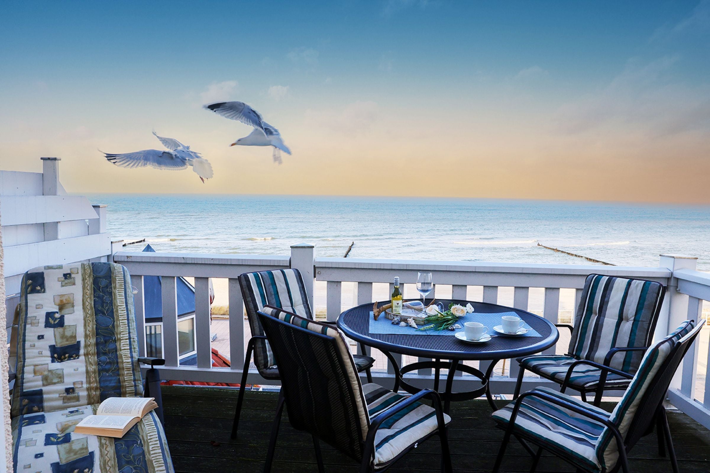 A terrace with a table and chairs, overlooking the sea, with seagulls flying in the background.