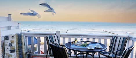A terrace with a table and chairs, overlooking the sea, with seagulls flying in the background.