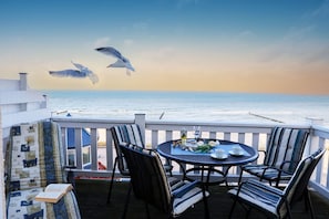 A terrace with a table and chairs, overlooking the sea, with seagulls flying in the background.