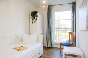 Quarto com cama, cadeira, mesa, planta pendente e janela com cortinas.