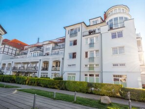 Um edifício residencial de vários andares, com varandas, uma torre de esquina e um jardim bem cuidado.