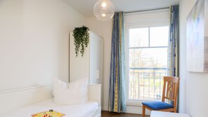 A bedroom with a bed, a chair, a small table, a hanging plant, and a window with curtains.