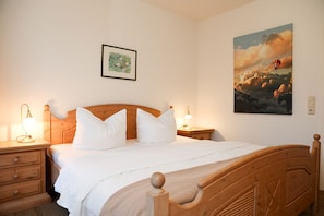 A bedroom with a wooden bed, white bedding, two bedside lamps, a nightstand, a drawer, and a framed picture on the wall.