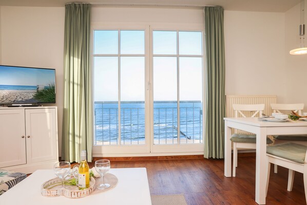 A room with a large window overlooking the sea, a dining table with chairs, a television on the left, and a white cabinet.