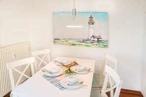 A dining table set with white plates, bowls, and cutlery, a painting of a lighthouse, and white chairs.