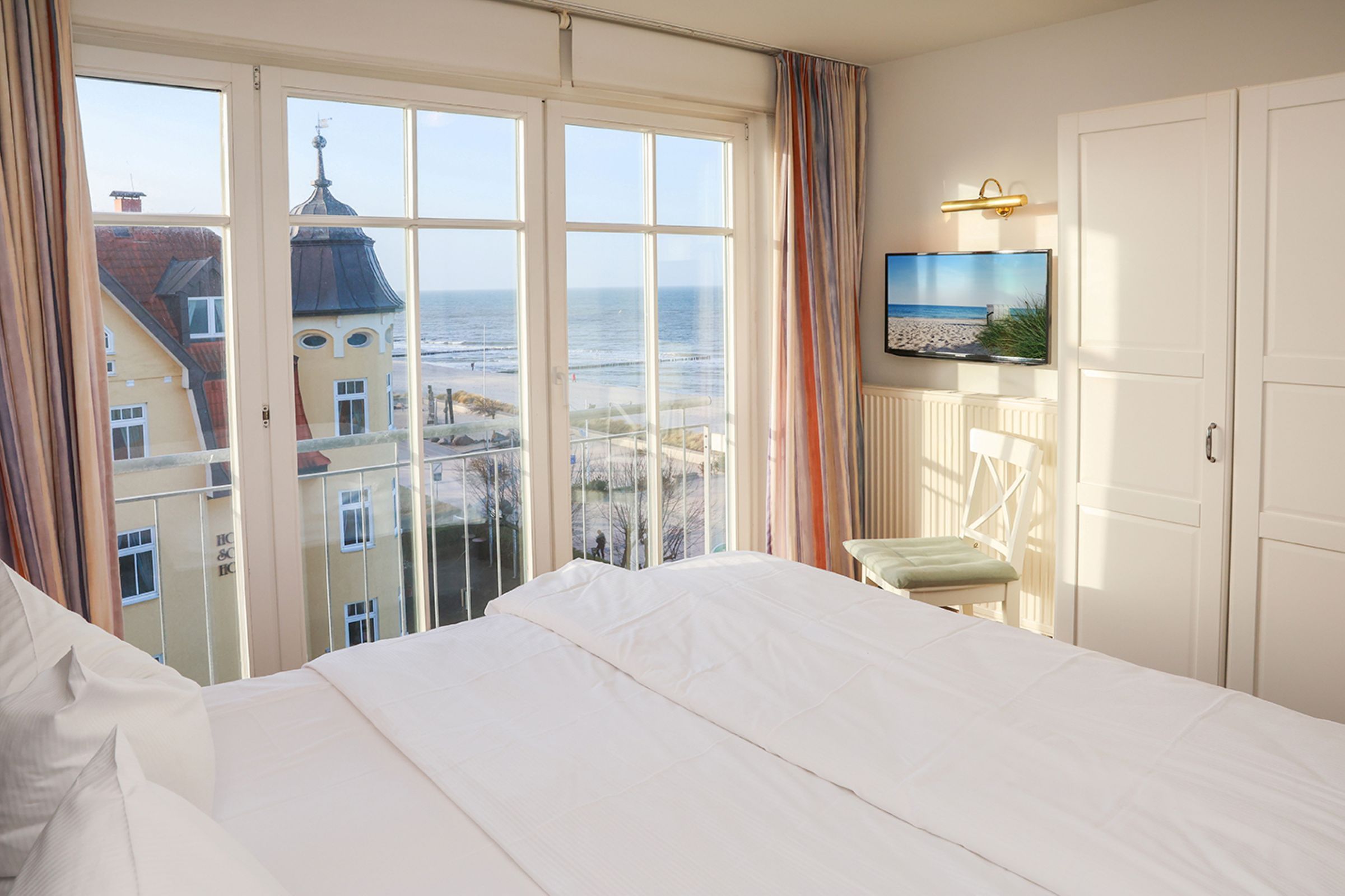 Quarto de hotel com uma cama grande, televisão na parede e vista para a praia e edifícios.