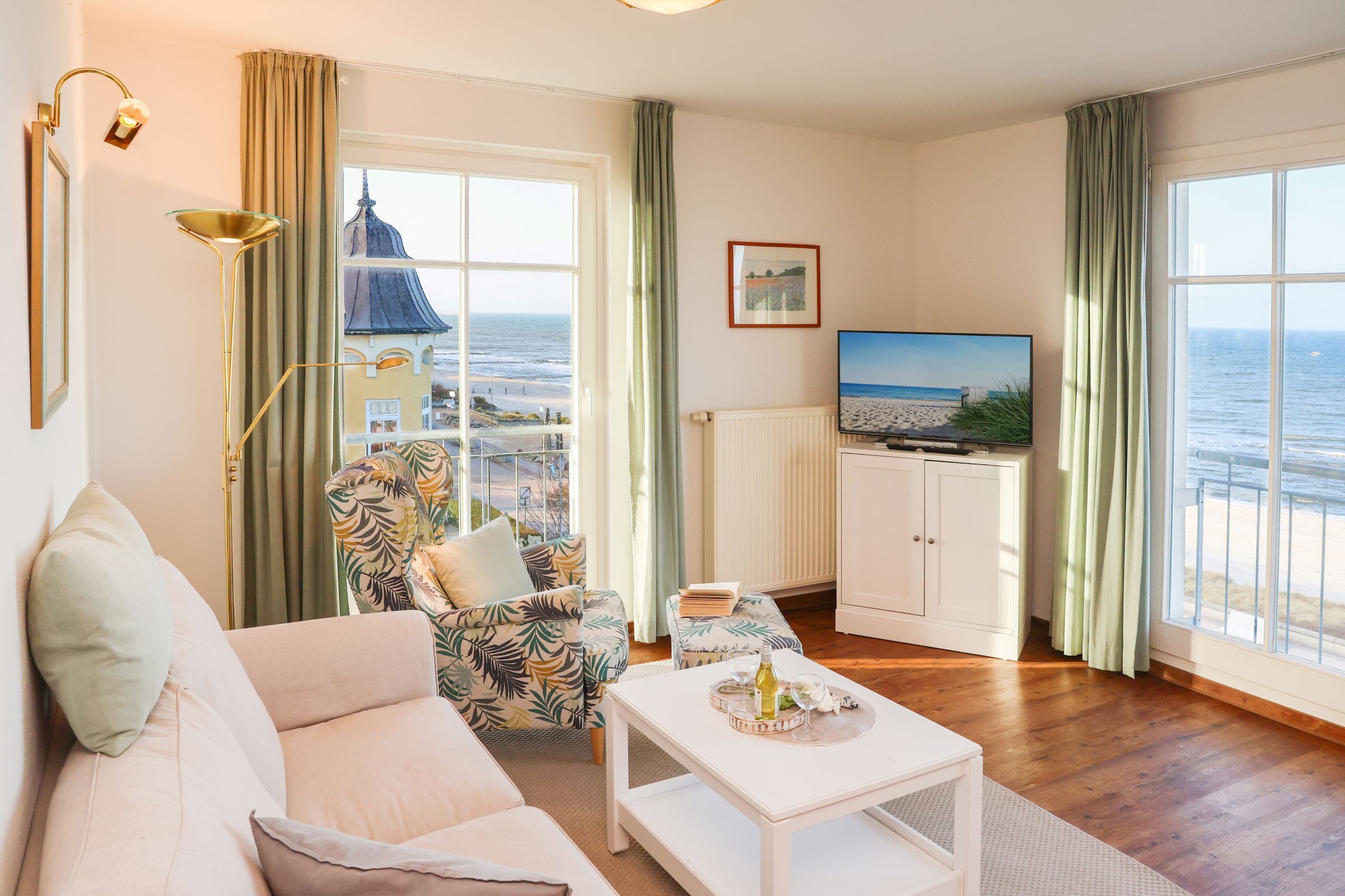 Un salon comprenant un canapé, un fauteuil à motifs, une petite table avec une bouteille et des verres, un téléviseur, et une vue sur la mer grâce à de grandes fenêtres.