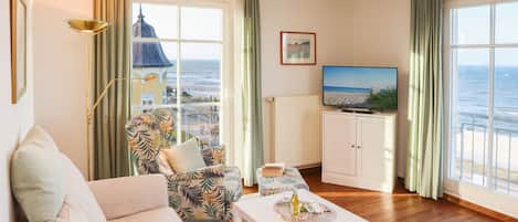 Una sala de estar con un sofá, un sillón estampado, una mesita con una botella y vasos, un televisor y vistas al mar a través de grandes ventanales.