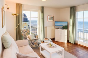 Sala de estar com sofá, poltrona estampada, mesa pequena com garrafa e copos, televisão e vista para o mar através de grandes janelas.