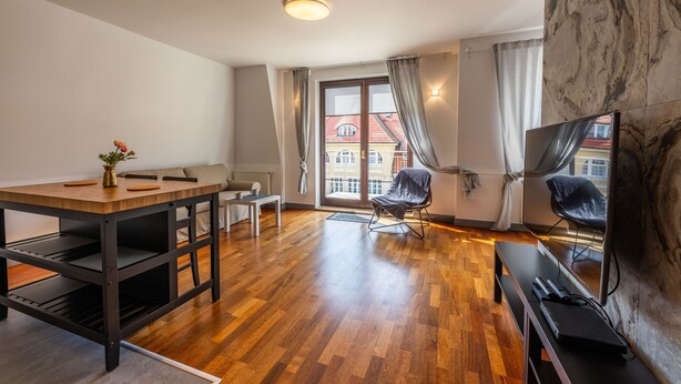 A modern living room with a wooden dining table, two chairs, a sofa, a TV, and a large window with curtains. - Victus Apartamenty - Prestige (Sopot)