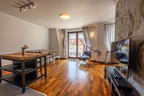 A modern living room with a wooden dining table, two chairs, a sofa, a TV, and a large window with curtains. - Victus Apartamenty - Prestige (Sopot)