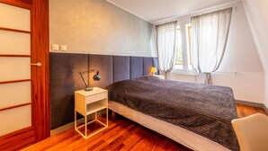 A modern bedroom with a wooden floor, a bed with a dark headboard, a bedside table with a lamp, and a window with curtains. - Victus Apartamenty - Prestige (Sopot)