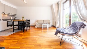 A modern kitchen with white cabinets, a dining table, and a sofa in a bright living area with wooden flooring and large windows. - Victus Apartamenty - Prestige (Sopot)