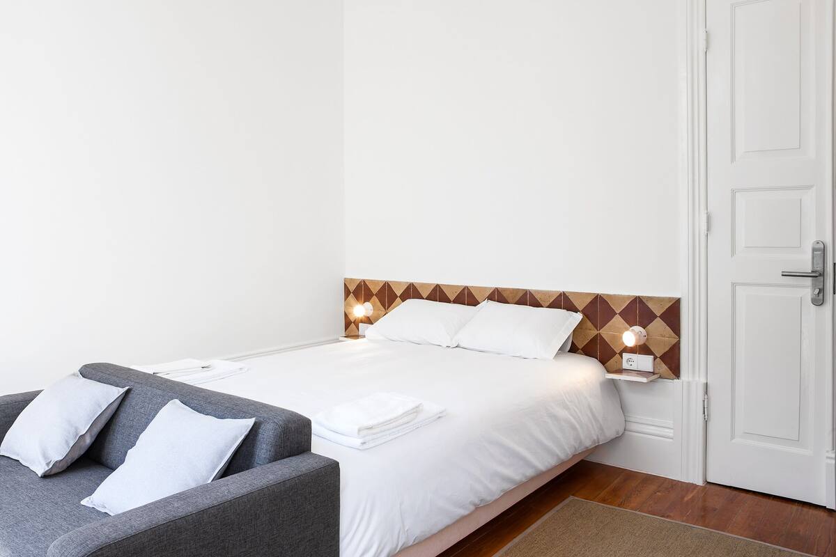 A bedroom with a bed, a grey sofa, a white door, and a patterned headboard.