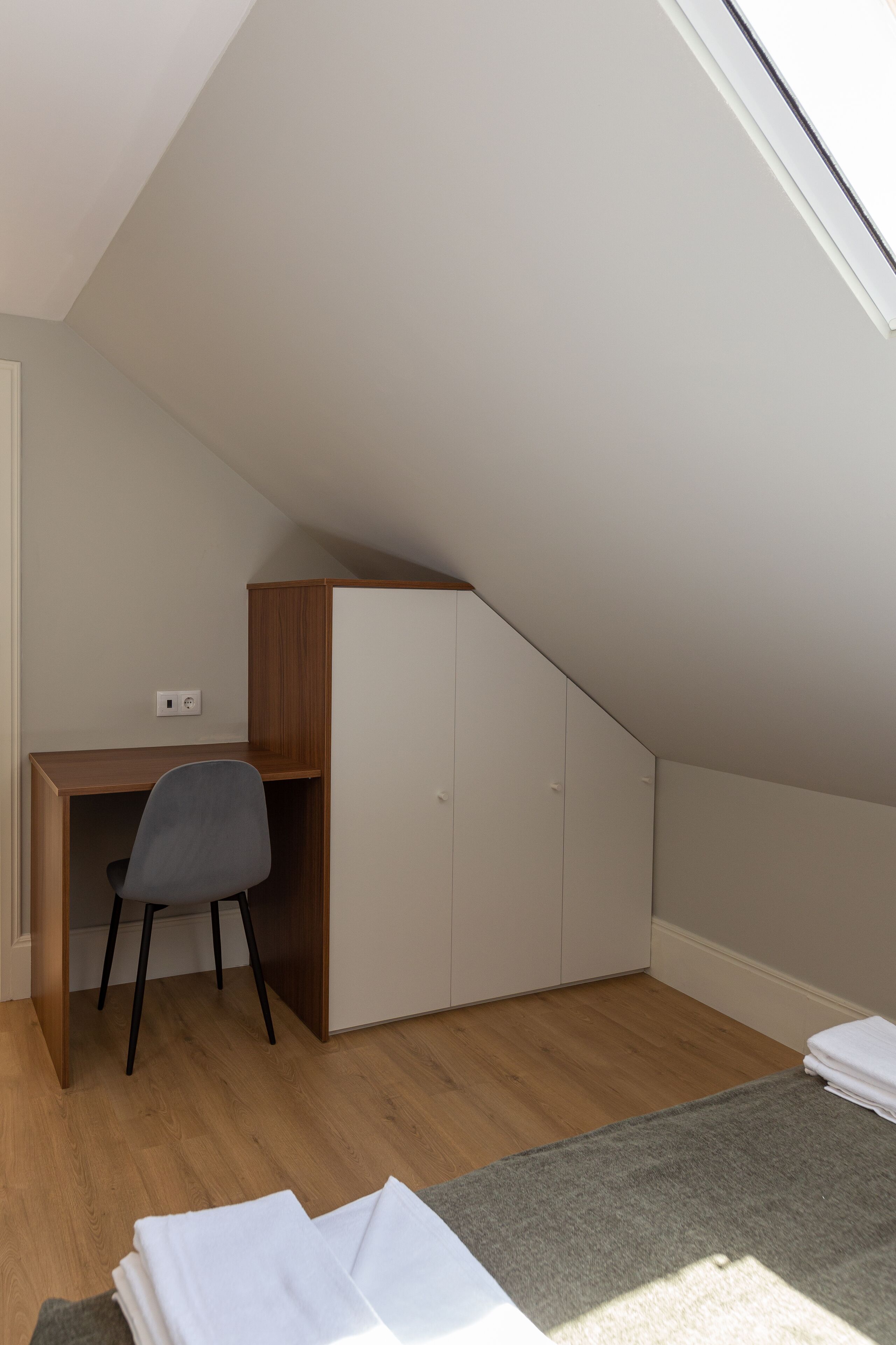 Une petite pièce moderne avec un plafond en pente, un bureau en bois, une chaise grise et une armoire blanche.