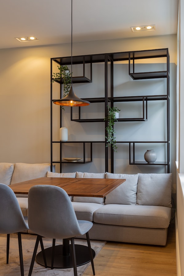 Uma sala de estar moderna com uma mesa de jantar de madeira, sofá revestido de cinza e estante de metal preto.
