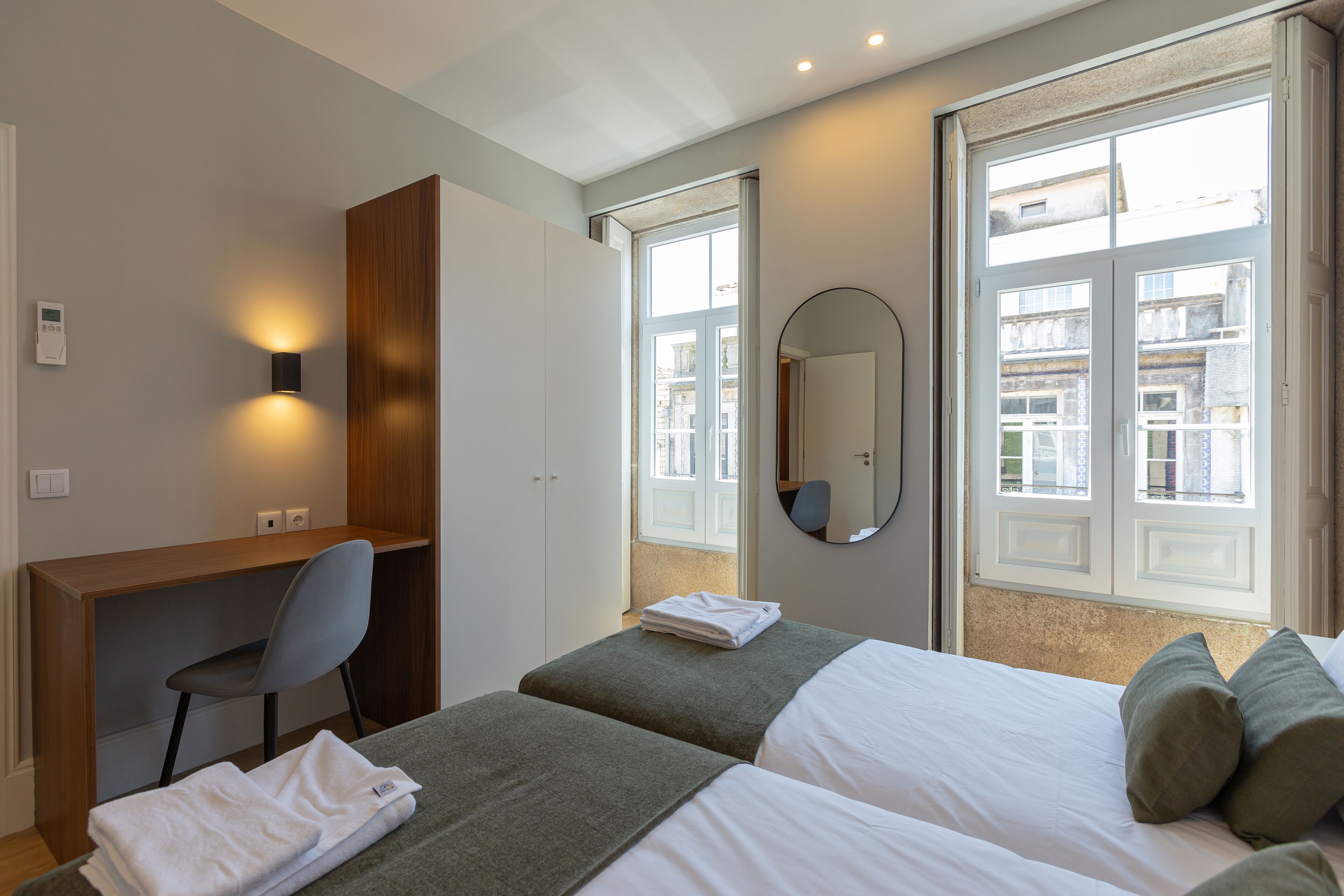 A hotel room with two beds, a desk, a chair, a mirror, and a window with a view of buildings.