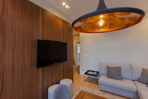 Uma sala de estar moderna com uma TV de tela plana fixada em uma parede de madeira, um sofá cinza claro e uma mesa redonda com uma revista.