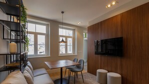 A modern living room with a sofa, a dining table with chairs, a TV mounted on the wall, and a staircase with a plant. - ORM - Bolhao Apartments (Porto)