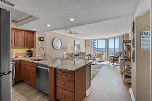 A kitchen with wooden cabinets, a marble countertop, and a dishwasher. There is a dining area with a table and chairs, and a view of the ocean through large windows.