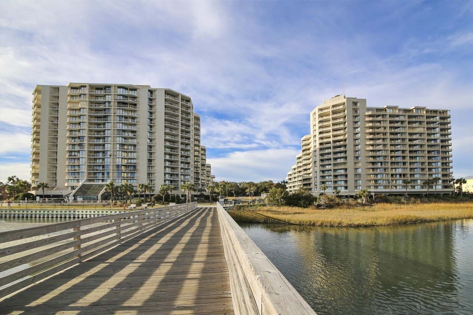 Two high-rise buildings with balconies, a wooden boardwalk, and a calm water body.
