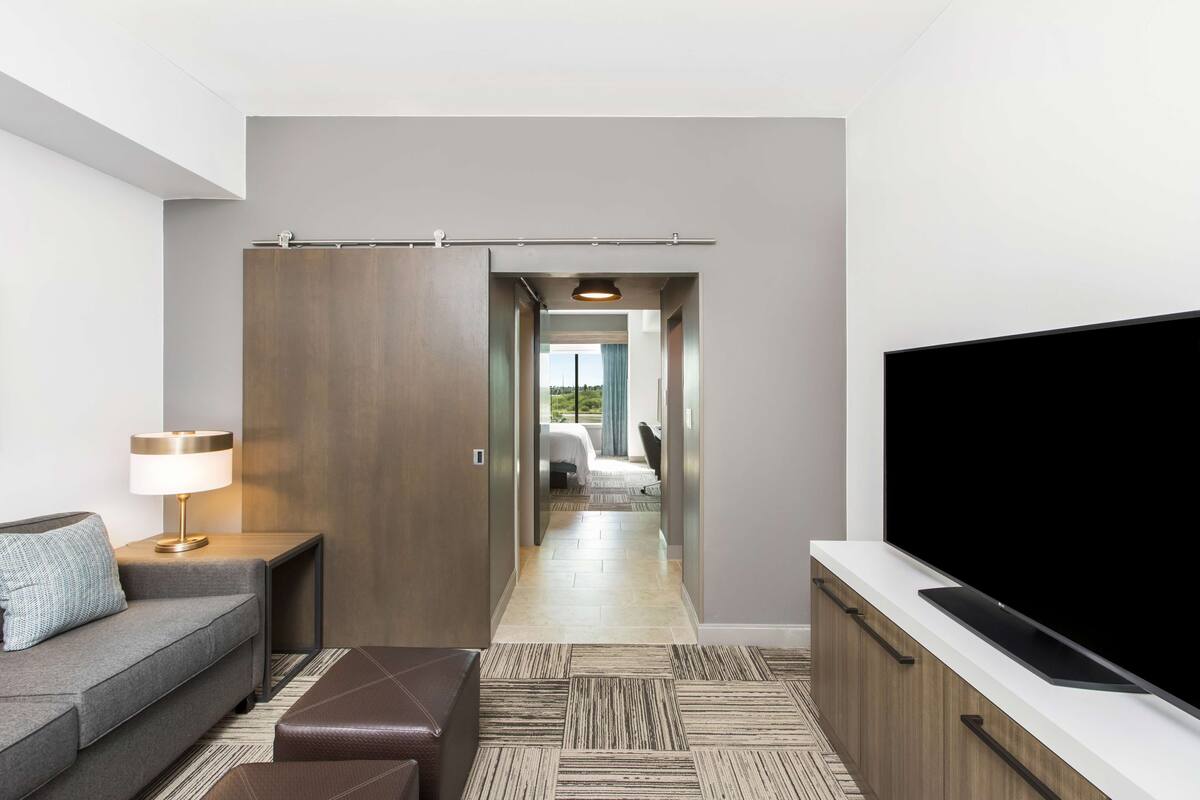 Ein modernes Hotelzimmer mit einer Couch, einem Fernseher, einer Lampe, einem Holzschrank und Blick ins Schlafzimmer.