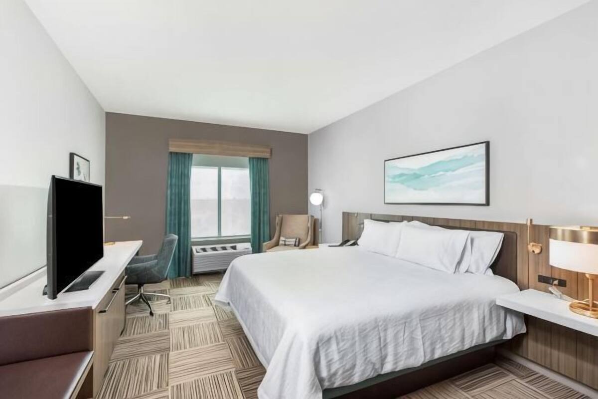 Ein Hotelzimmer mit Bett, Schreibtisch, Stuhl, Fernseher und einem Fenster mit Vorhängen.