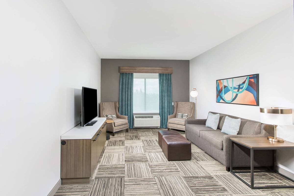 Ein modernes Hotelzimmer mit Flachbildfernseher, zwei Sesseln, einem Sofa, einem Couchtisch und einem großen Fenster mit Vorhängen.
