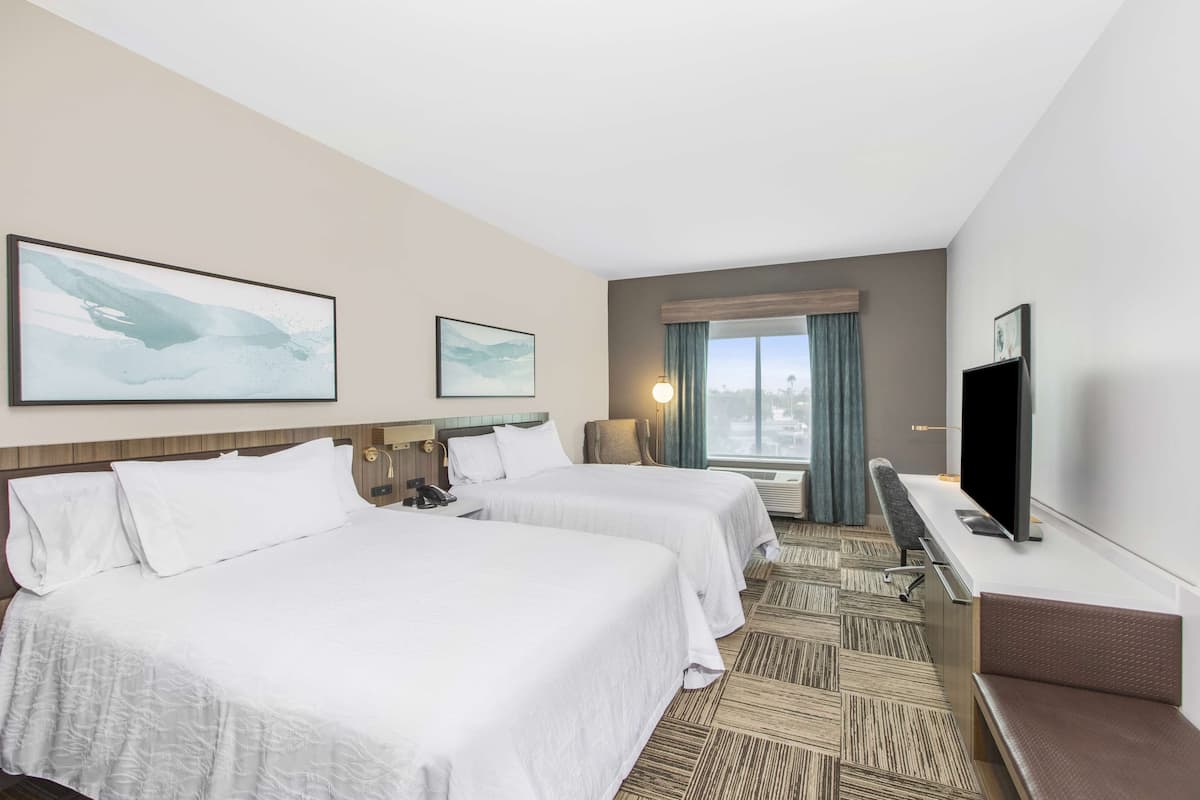Ein Hotelzimmer mit zwei Betten, einem Schreibtisch mit Fernseher und einem Fenster mit Vorhängen.