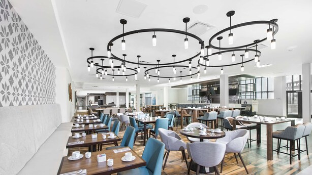 Un espace de restauration moderne dans un hôtel, avec de nombreuses tables et chaises, un mur décoré de motifs et un grand lustre.