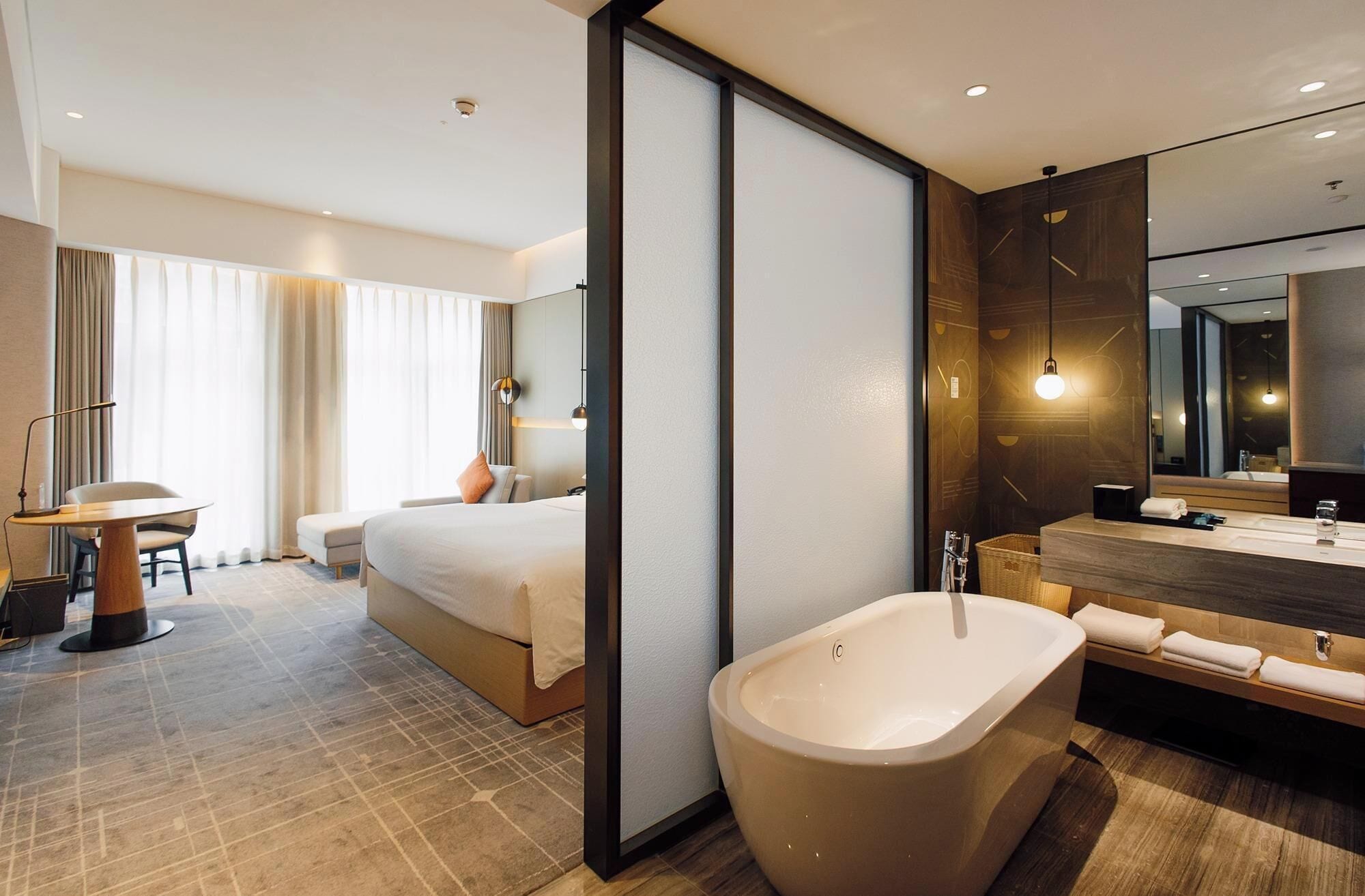 Ein modernes Hotelzimmer mit einem großen Bett, einer separaten Sitzecke mit Tisch und Stuhl, einer großzügigen Badewanne, einem Schminktisch mit Spiegel und einem großen Fenster mit Vorhängen.