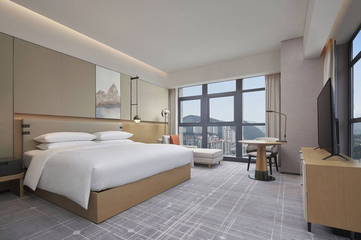 A modern hotel room with a large bed, a desk, a chair, and a large window offering a city view.